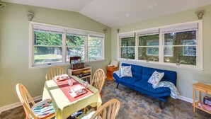Una habitación acogedora con una mesa de comedor, sillas, un sofá azul y ventanas amplias que dan a vistas de vegetación.