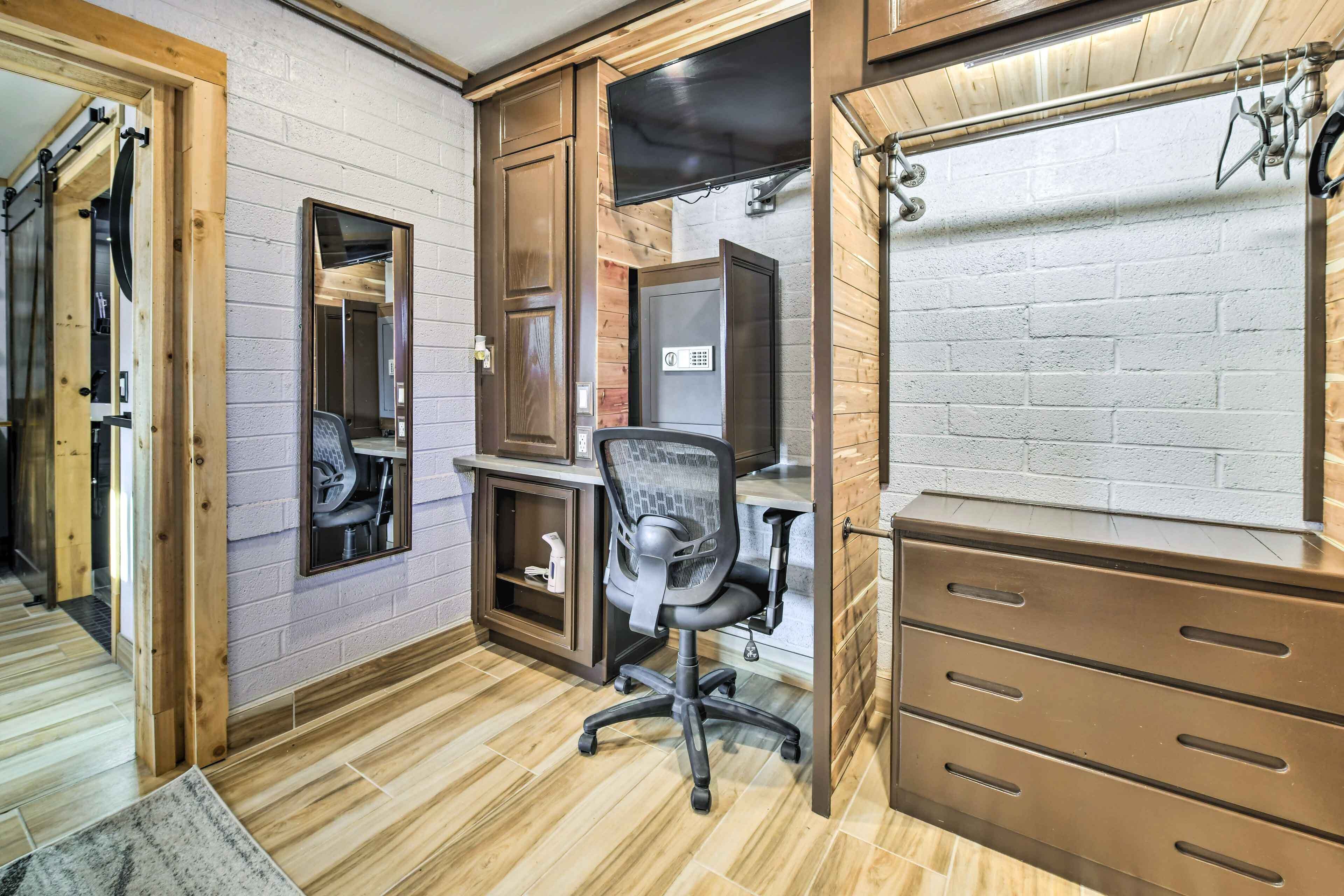 A room with a wooden desk, a chair, a television mounted on the wall, and a wardrobe with drawers.