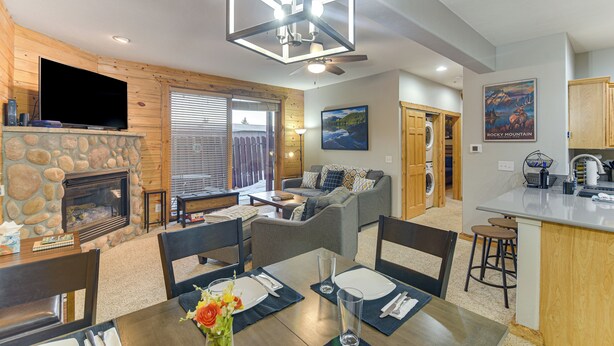A cozy living area with a stone fireplace, a flat-screen TV, a dining table set for four, and a kitchen with a bar area. - Patio & Mountain Views: Central Grand Lake Condo! (Grand Lake)