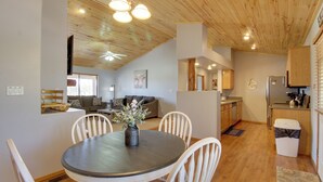 Eine moderne Küche mit Holzschränken, ein Essbereich mit rundem Tisch und Stühlen sowie ein Wohnzimmer mit Sofa und Deckenventilator.