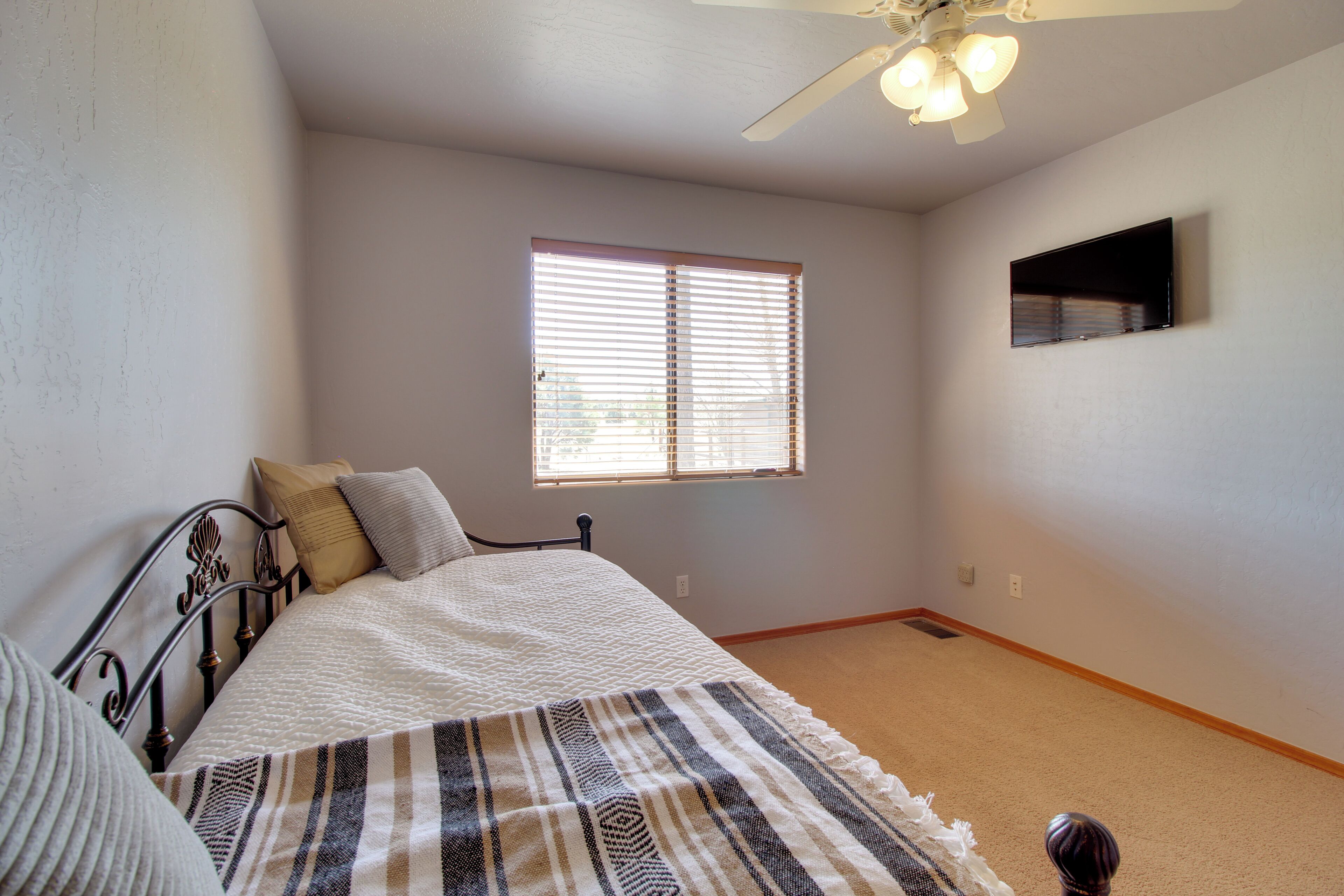 A bedroom with a bed, a window with blinds, a ceiling fan, and a wall-mounted TV.