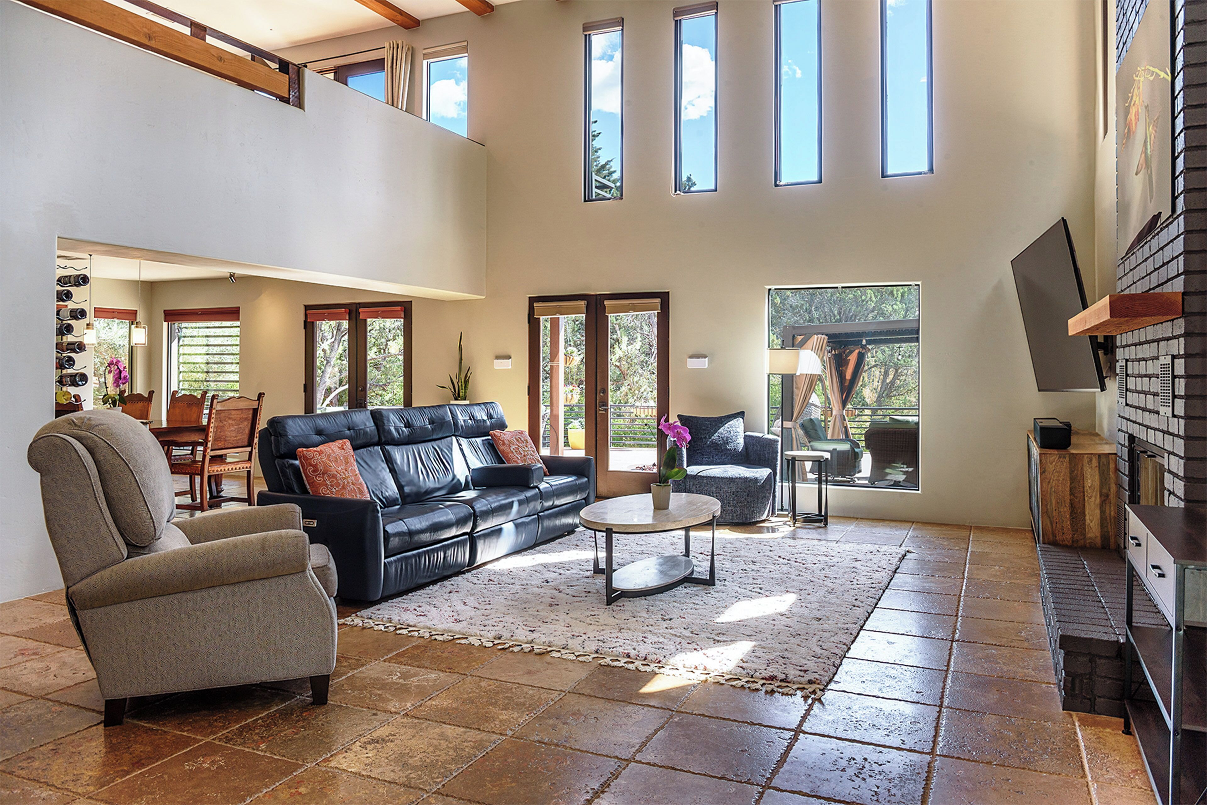 Amplia sala de estar con techo alto, un sofá de cuero negro, un sillón gris y una mesita de centro. Varias ventanas ofrecen luz natural y vistas al exterior.