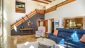 Un vaste espace de vie avec un canapé bleu, un escalier en bois et une cuisine visible à travers une porte ouverte.