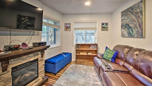 A living room with a stone fireplace, a flat-screen TV, a leather sofa, a blue ottoman, and a bookshelf. - 1st-floor Wintergreen Resort Condo w/ Balcony (Nellysford)