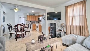 A living room with a sofa, a dining table with chairs, a TV, and a kitchen in the background.