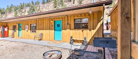 A wooden cabin with a blue door, a fire pit, and two chairs.