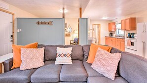 A cozy living room with a grey sofa, orange and patterned cushions, and a kitchen in the background.