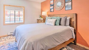 A bedroom with a large bed, a ceiling fan, a window with blinds, and a wall with framed pictures.