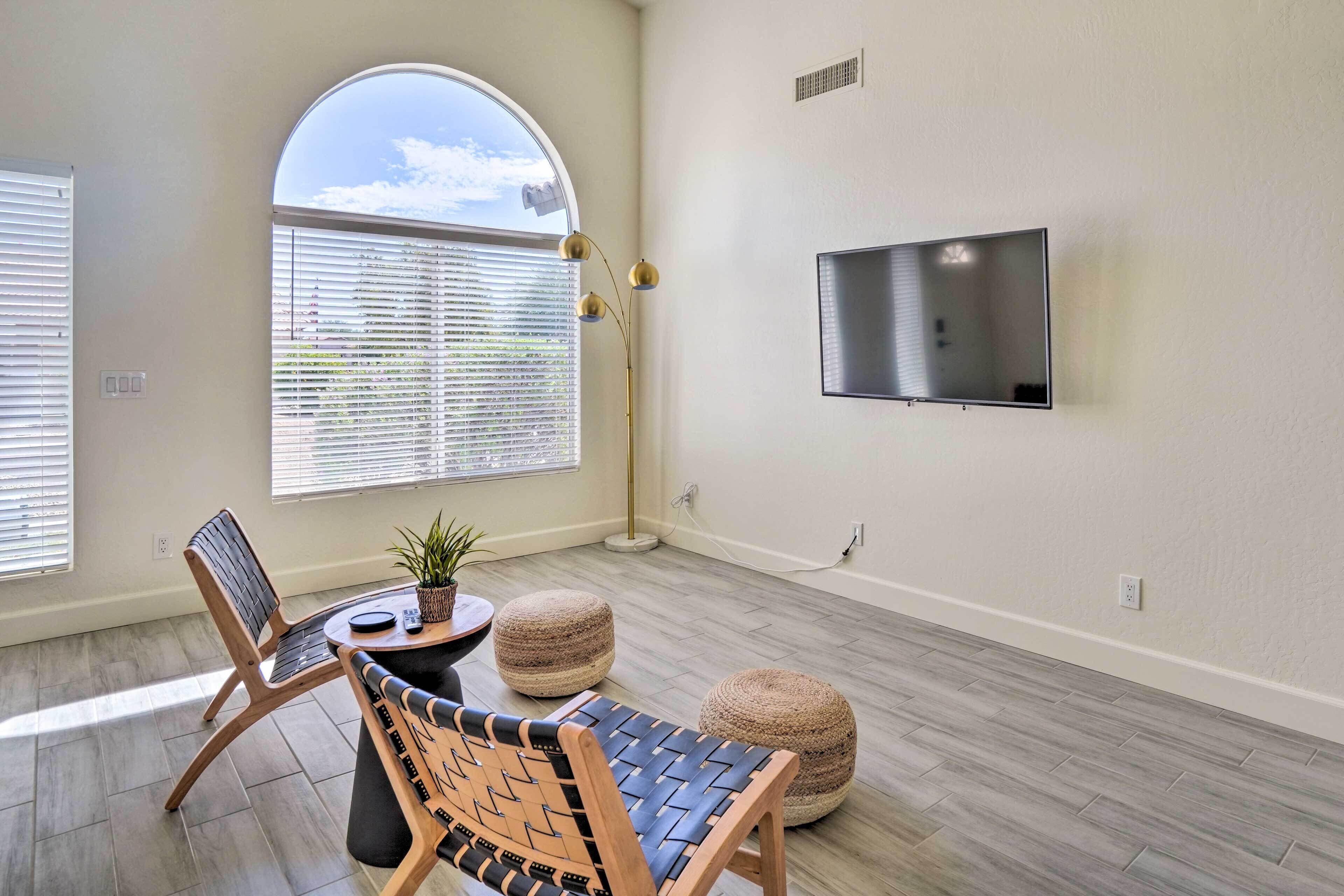 A modern living room with a large window, a flat-screen TV mounted on the wall, a wooden chair, a round ottoman, and a floor lamp.