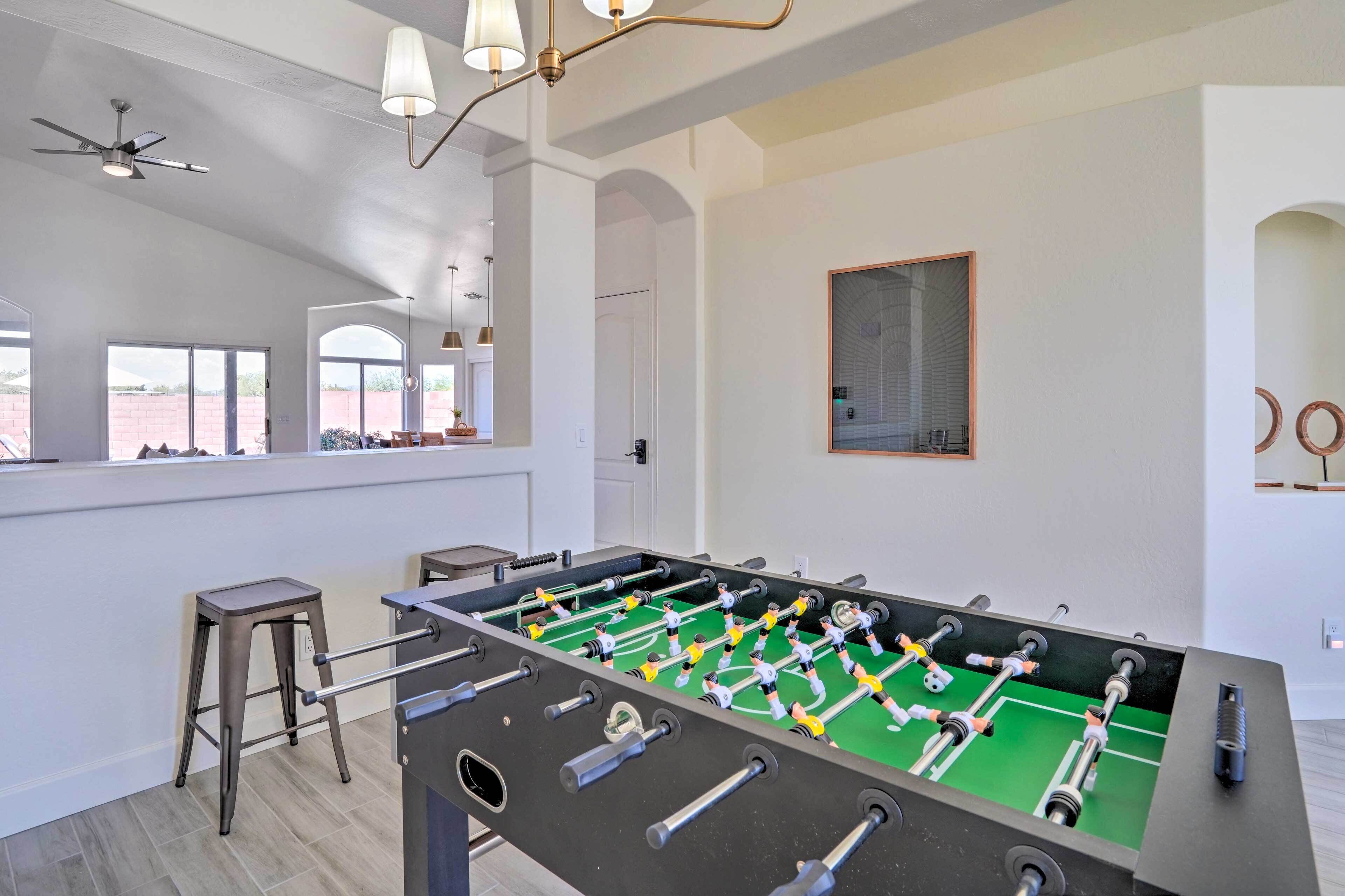 A modern room with a foosball table, two bar stools, a ceiling fan, and a large window with a view of the outside.