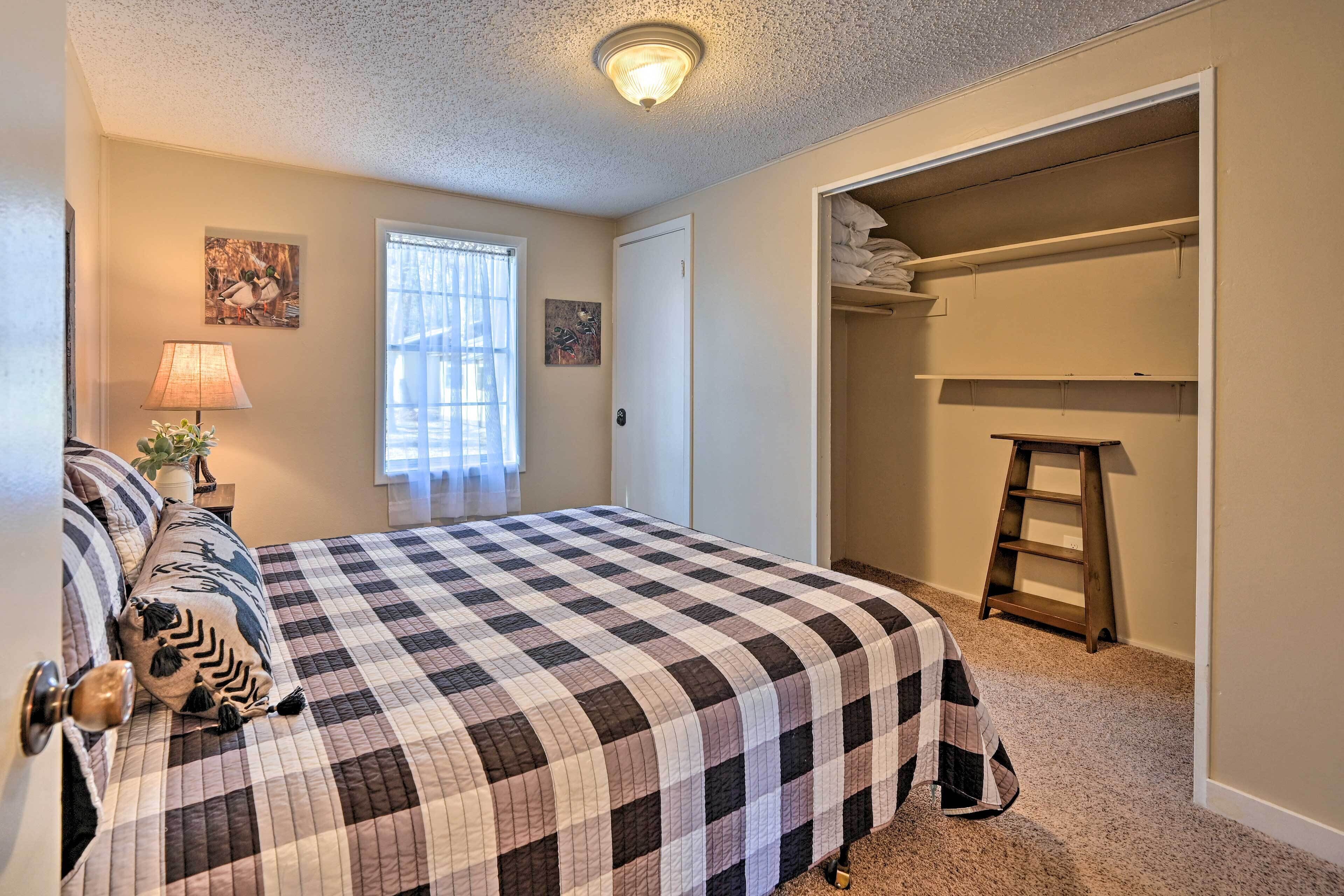 A bedroom with a checkered bedspread, a bedside lamp, a window with curtains, a door, and a closet with shelves.