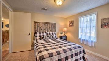 A bedroom with a checkered bedspread, a wooden headboard, a bedside lamp, and a window with sheer curtains.