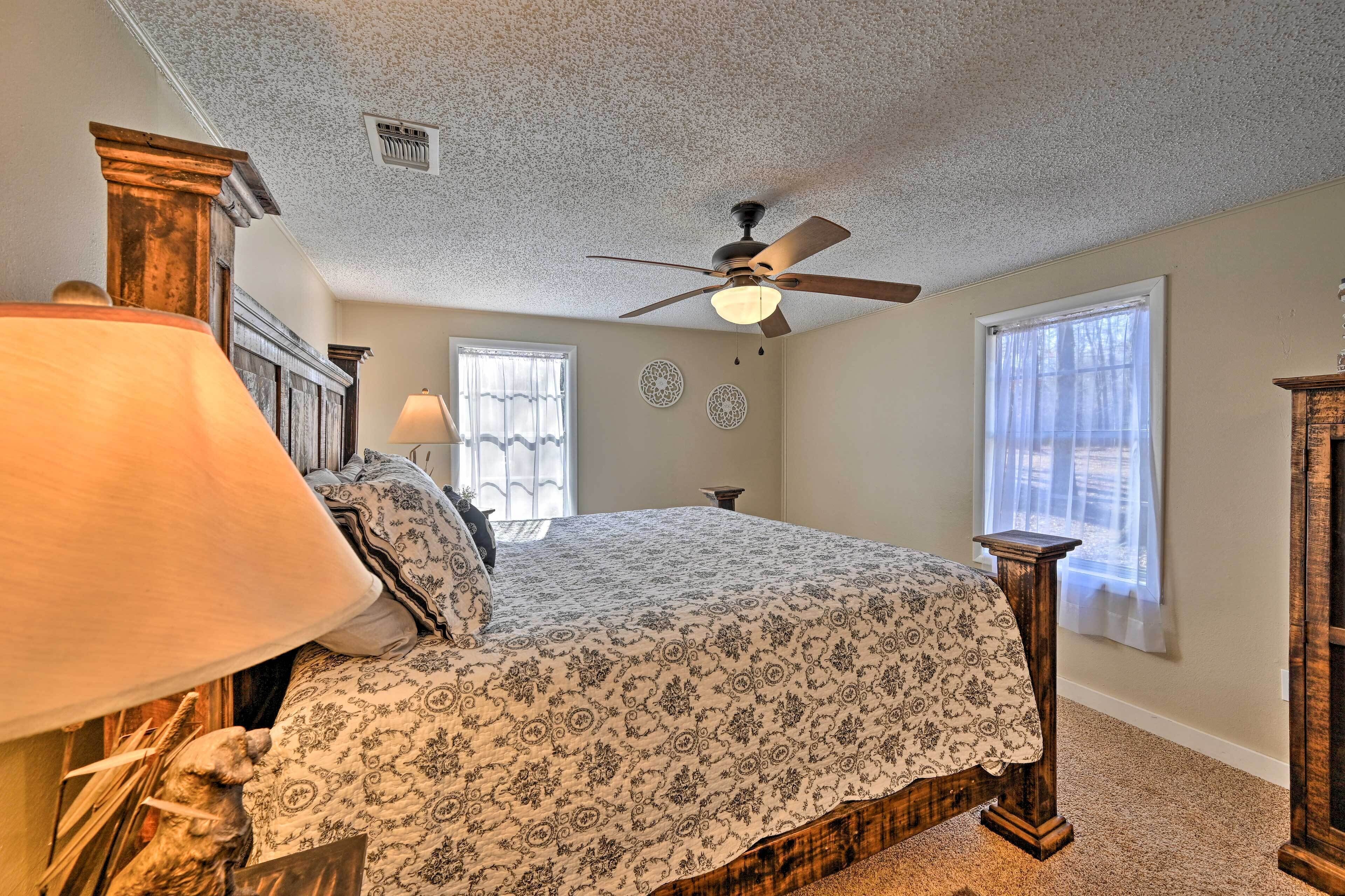 A bedroom with a bed, a ceiling fan, two windows, and a lamp.