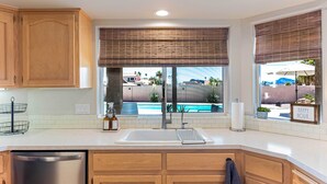 A kitchen with wooden cabinets, a stainless steel dishwasher, a white sink, and a view of a swimming pool.