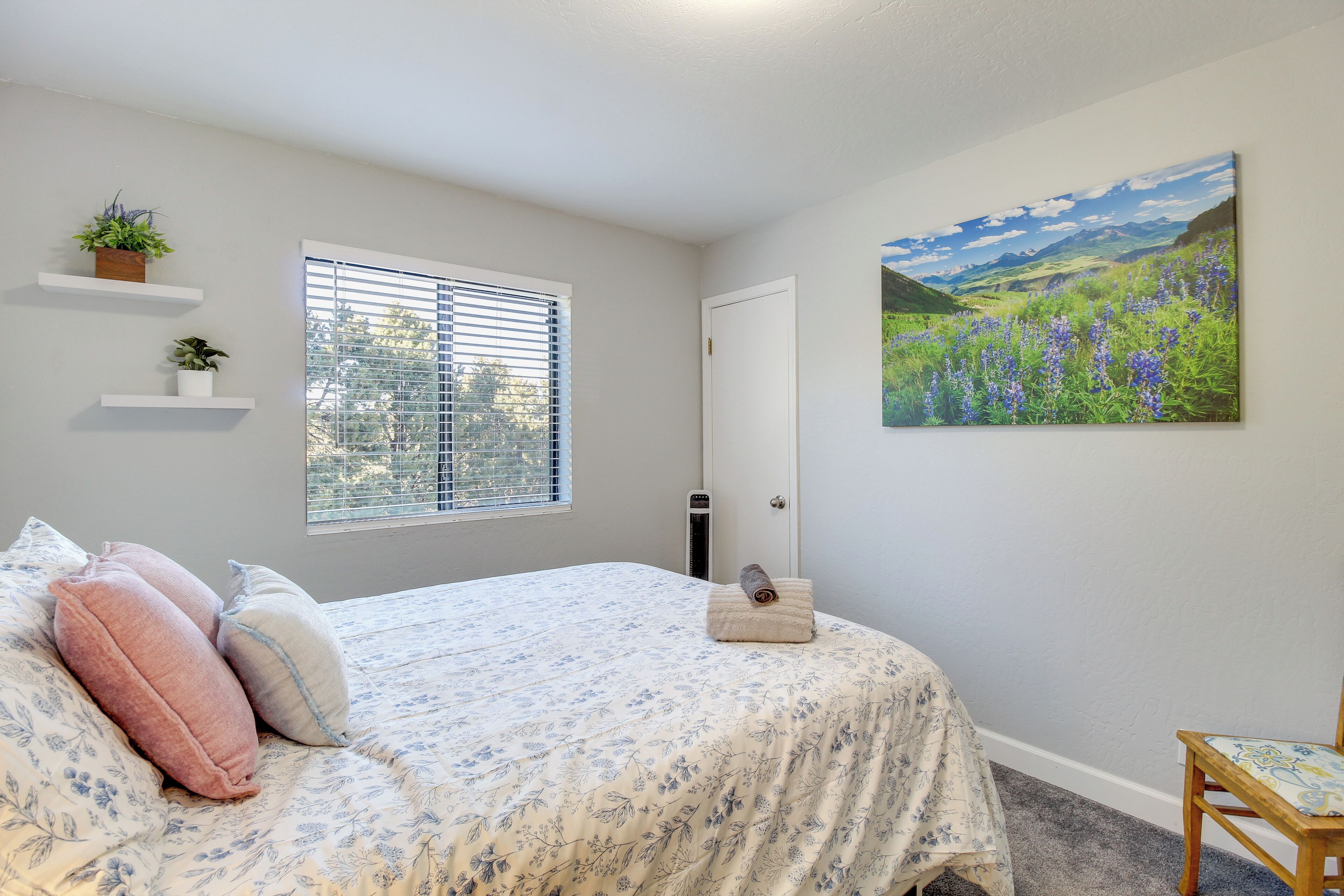 Un dormitorio con cama, ventana con persianas, puerta, estante montado en la pared con plantas y un cuadro de un paisaje.
