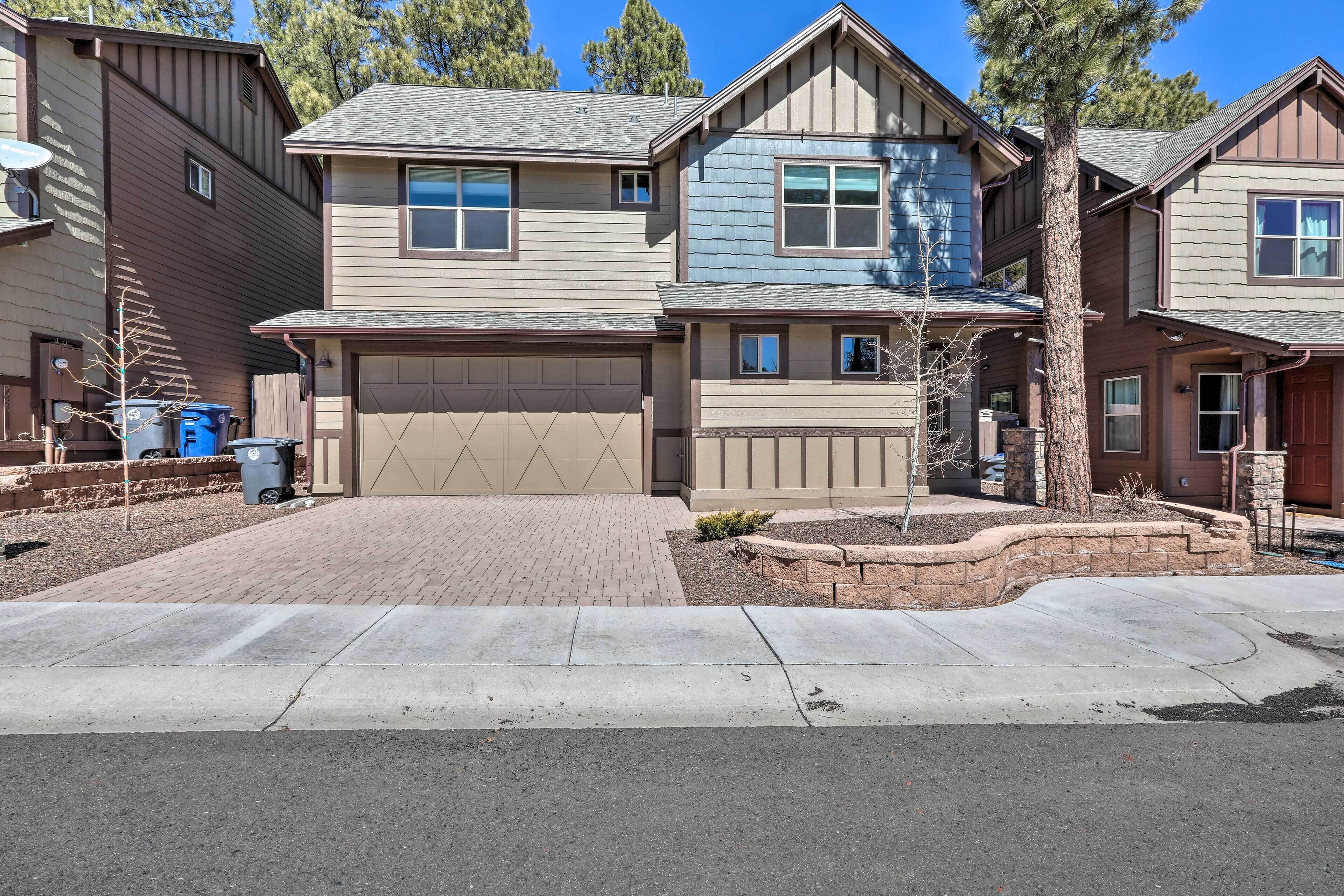A two-story residential building with a beige garage door, a brick driveway, and a landscaped front yard with a tree and a small bush.