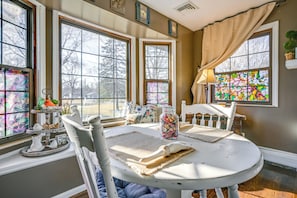 A dining area with a round table, white chairs, and a view of trees through large windows.