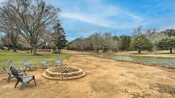 A circular fire pit with chairs around it, set in a sandy area with trees and a body of water in the background.