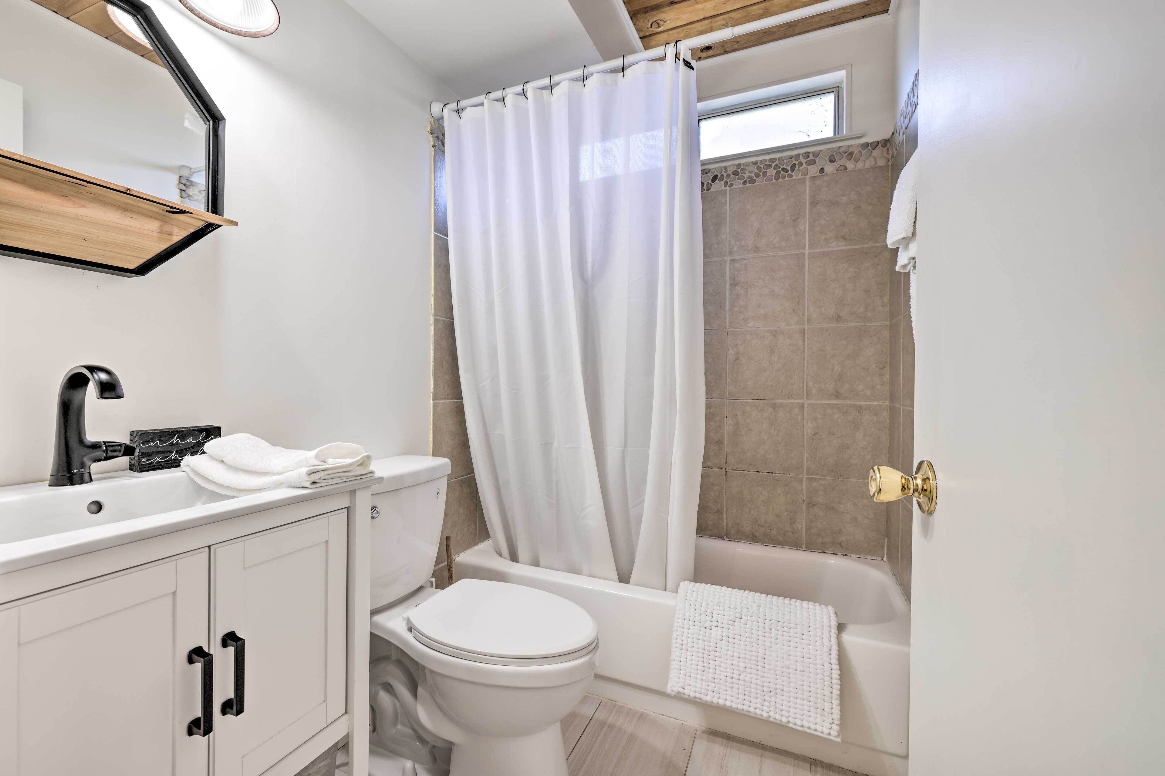 Un baño moderno con lavamanos, inodoro y bañera blancos. Se ve una ventana sobre la bañera, y un perchero.