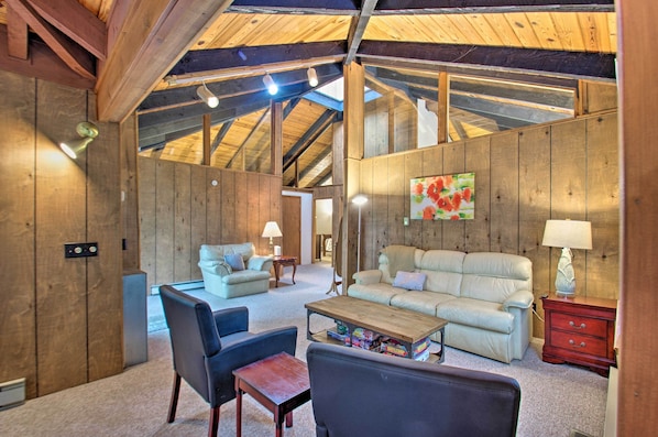 A spacious living room with a high ceiling, wooden walls, and a cozy fireplace. There are two sofas, a coffee table, and a TV.
