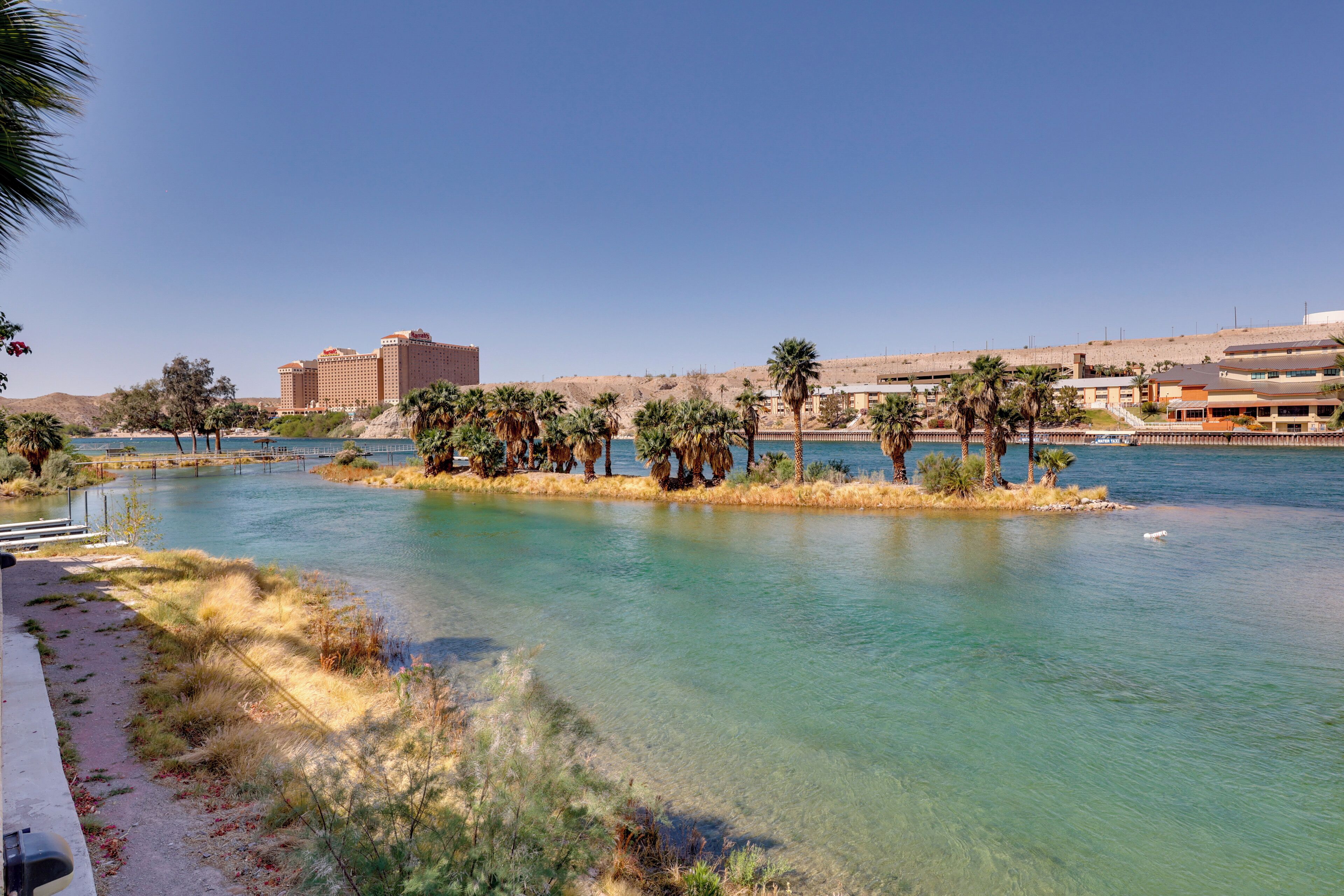 A river with clear water, surrounded by greenery and palm trees. There are buildings in the background.