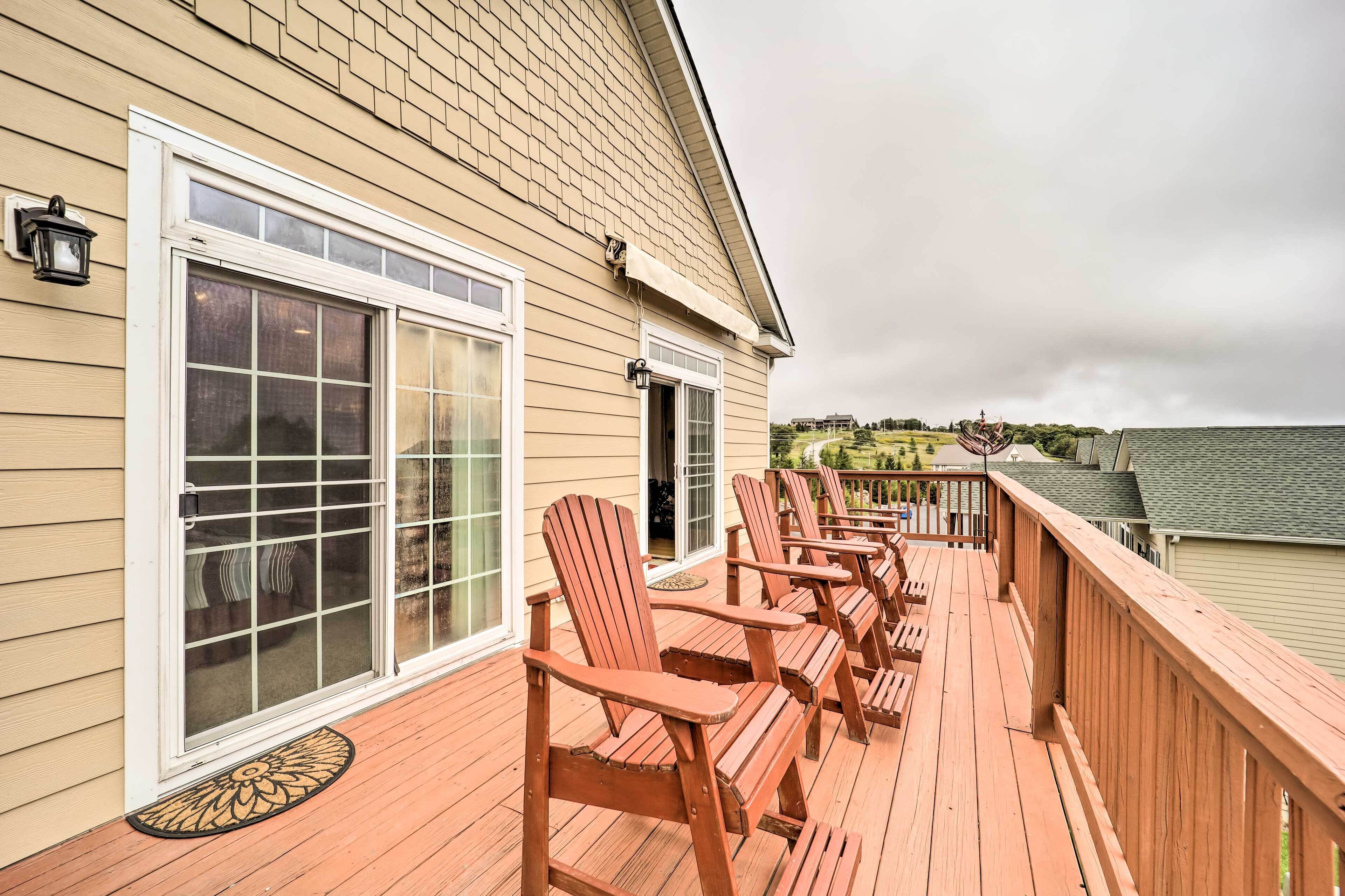 A wooden deck with Adirondack chairs, a covered area, and a view of a green house in the background.