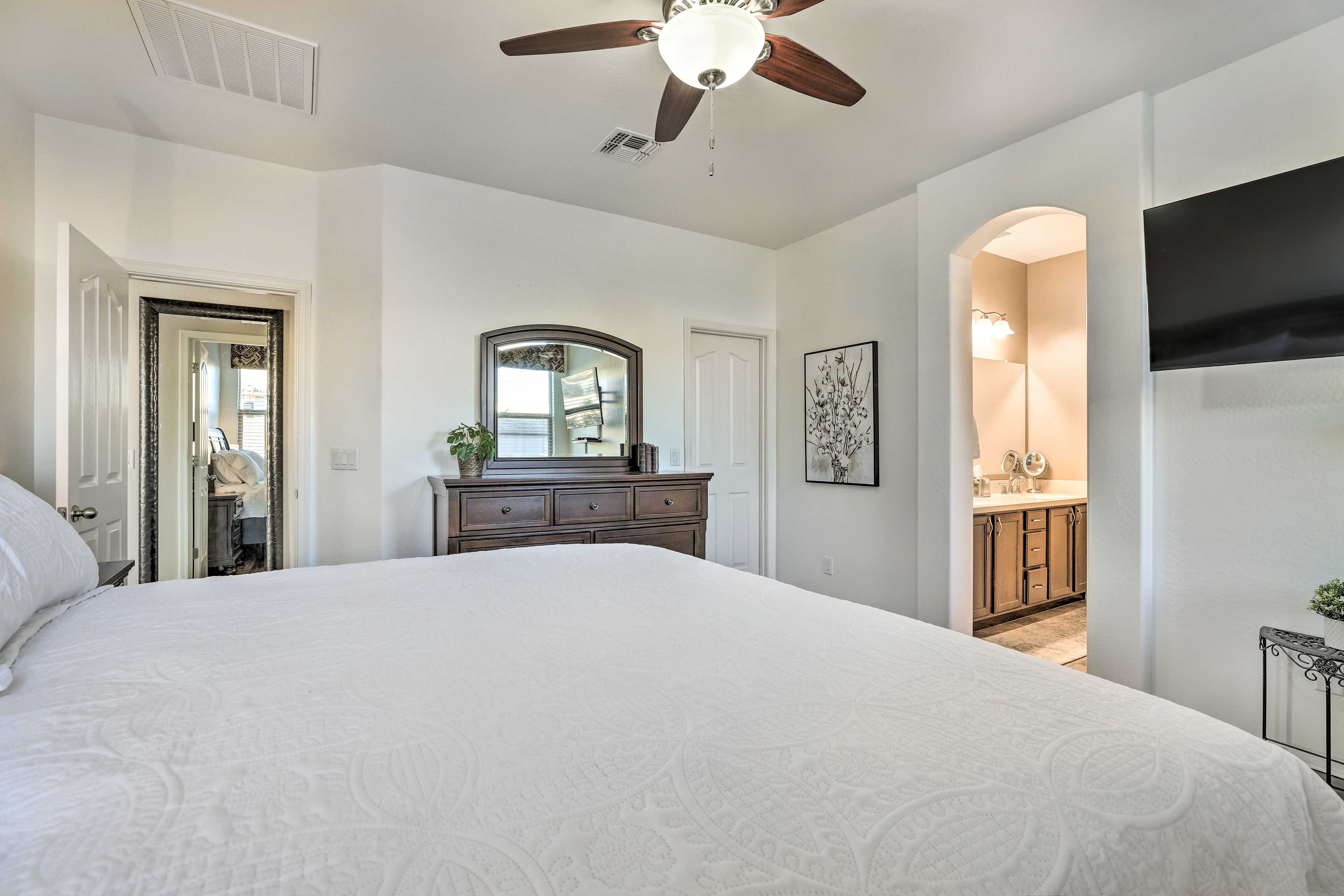 A bedroom with a large bed, a ceiling fan, a dresser, a mirror, and a TV mounted on the wall.
