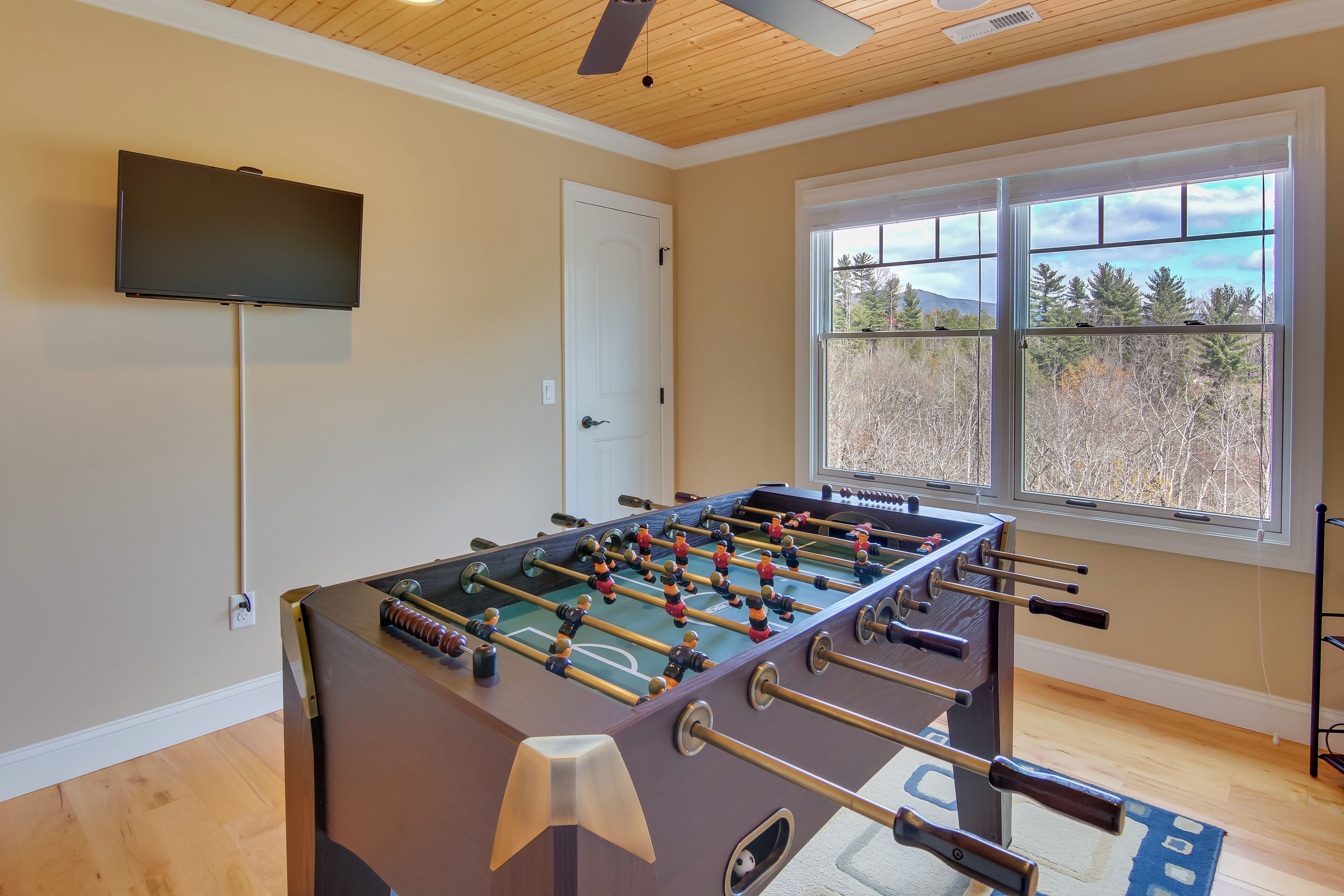 A room with a foosball table, a television mounted on the wall, and a window with a view of trees and mountains.