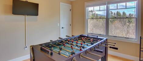 A room with a foosball table, a television mounted on the wall, and a window with a view of trees and mountains.