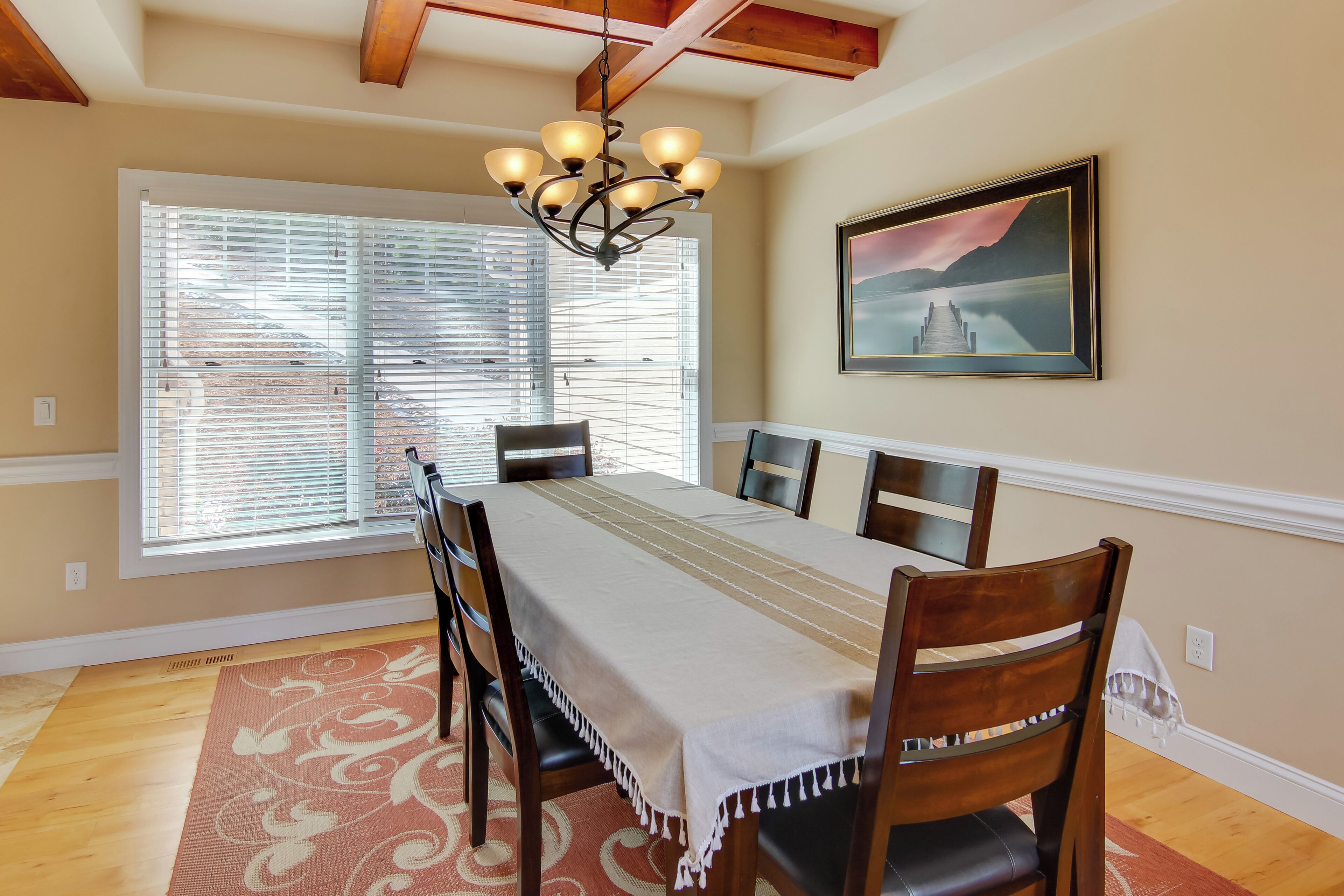 A dining room with a long table, wooden chairs, a chandelier, a painting, and a window with blinds.