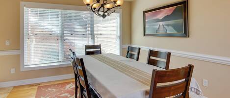 A dining room with a long table, wooden chairs, a chandelier, a painting, and a window with blinds.