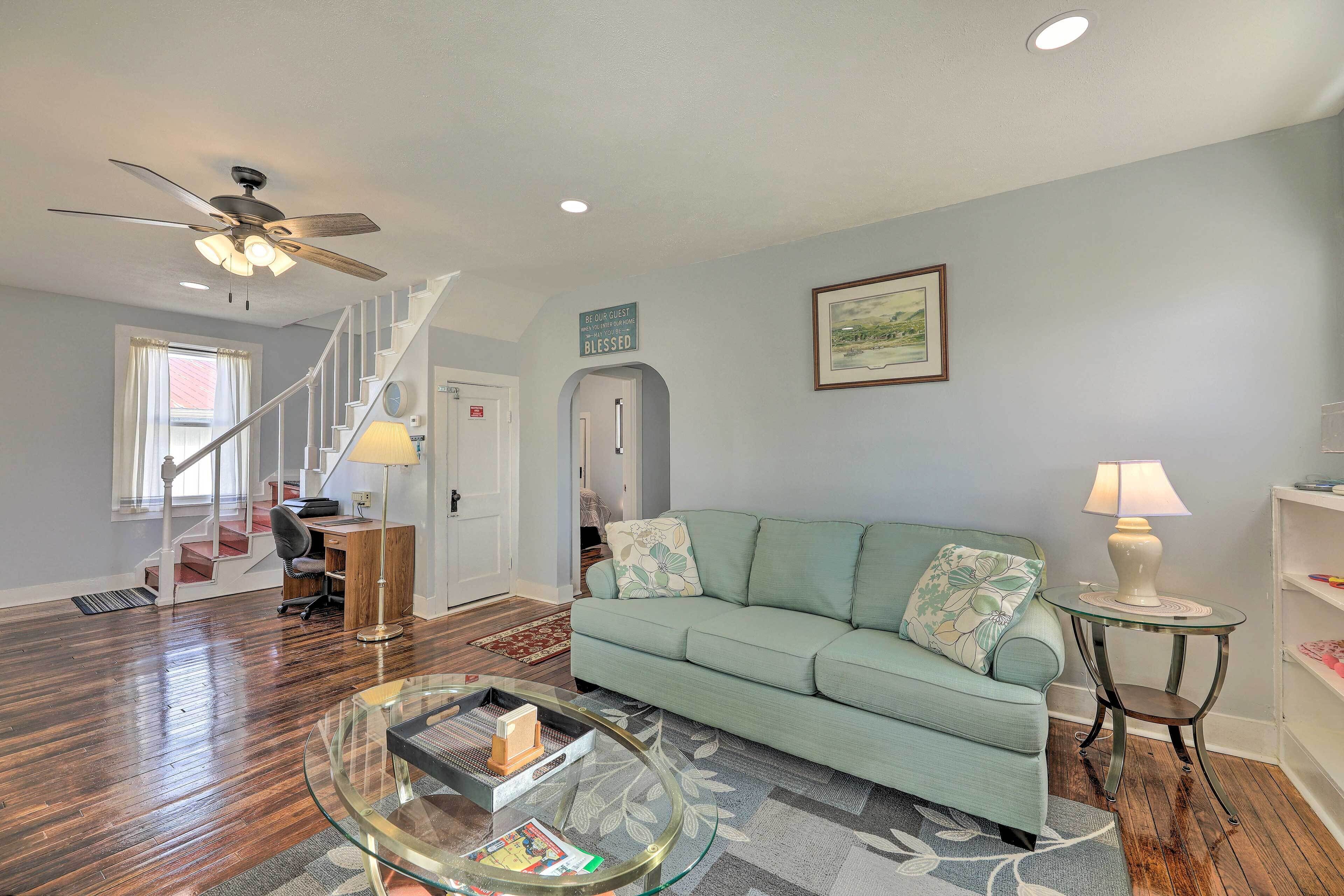 A living room with a green sofa, a glass coffee table, a ceiling fan, and a staircase leading to another room.