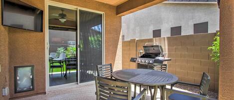An outdoor patio with a round table and chairs, a ceiling fan, a TV mounted on the wall, and a grill.