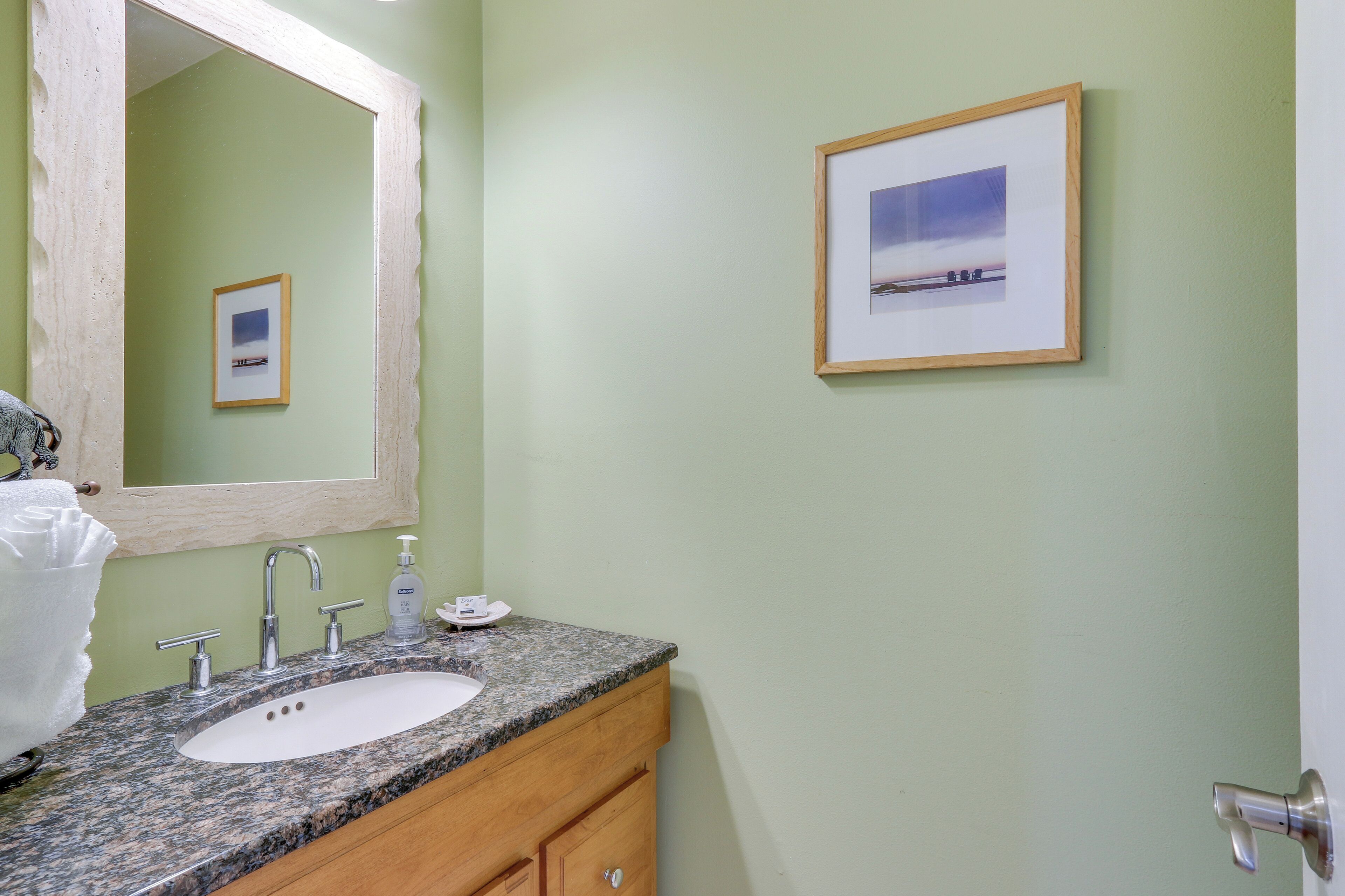 A bathroom with a granite countertop, a white sink, a wooden vanity, a large mirror, and a framed picture on the wall.