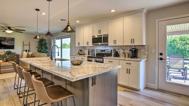 A modern kitchen with white cabinets, stainless steel appliances, a large island with bar stools, and a view to the outdoors. - Putting Green, Community Perks: Sun City West Home (Sun City West)