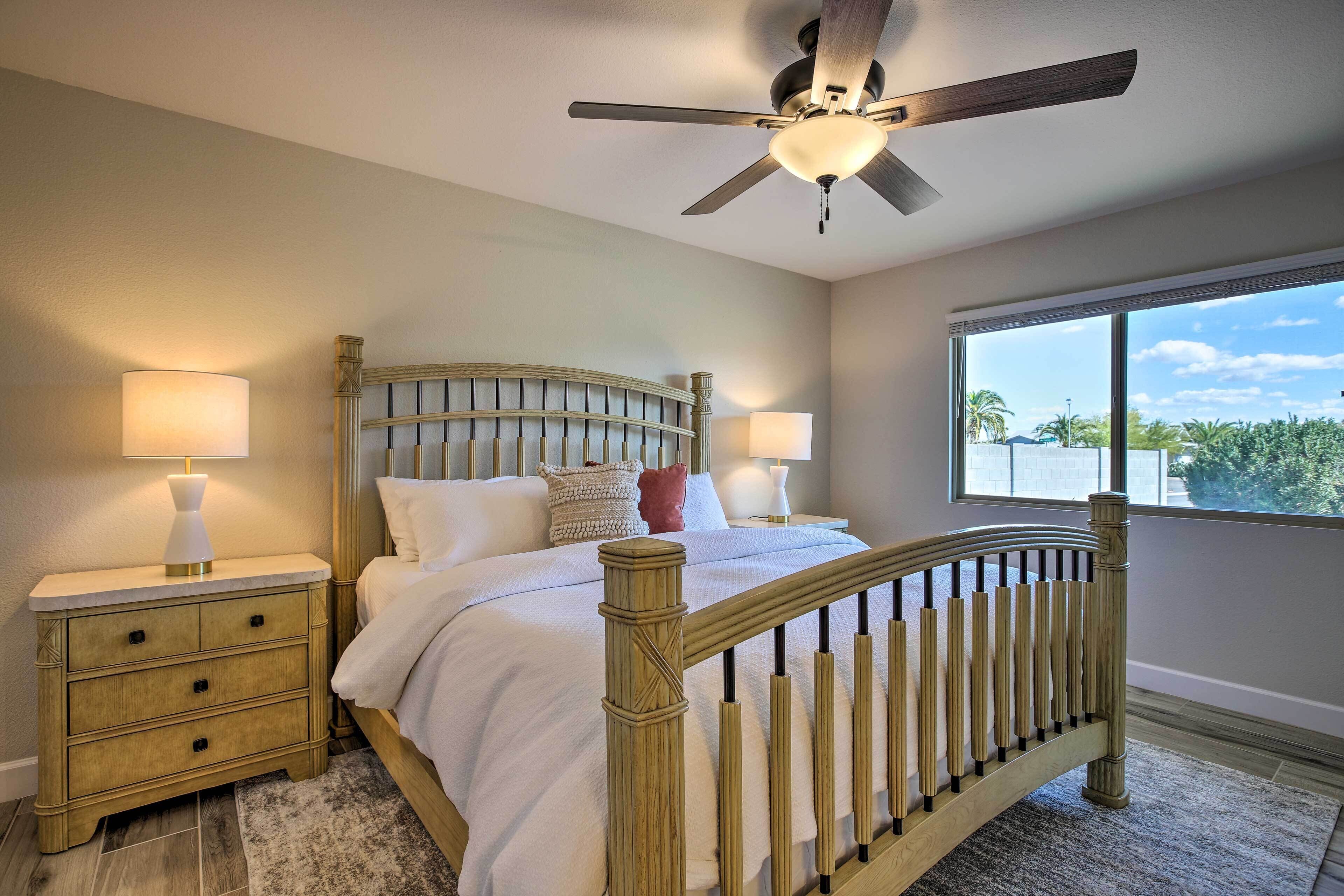A bedroom with a wooden bed frame, two bedside tables with lamps, a ceiling fan, and a large window with a view of palm trees.