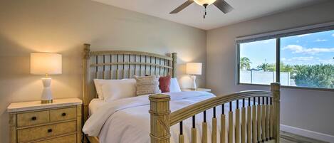A bedroom with a wooden bed frame, two bedside tables with lamps, a ceiling fan, and a large window with a view of palm trees.