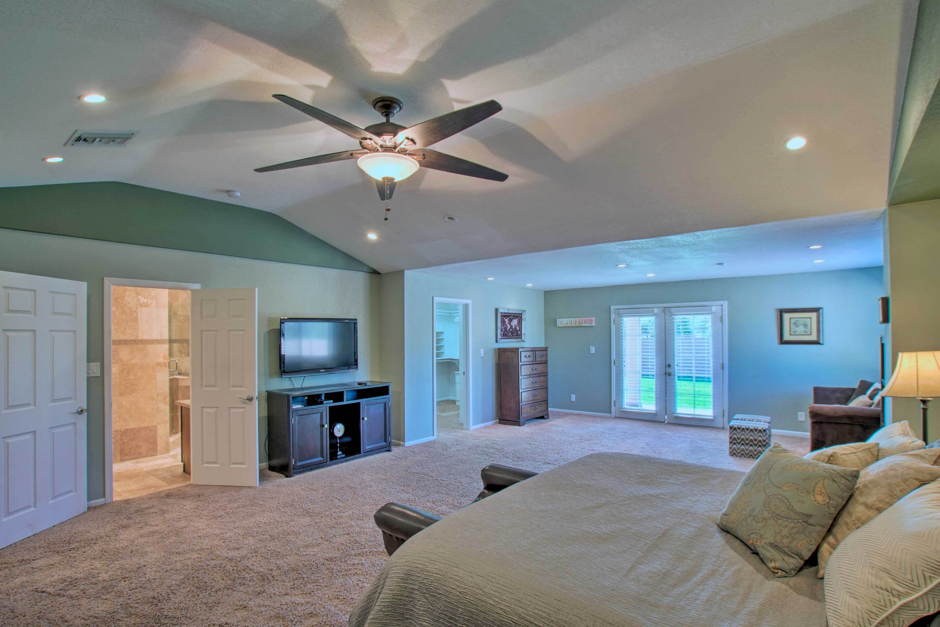 A spacious bedroom with a large bed, a ceiling fan, a television, and a view of the outside through a sliding glass door.
