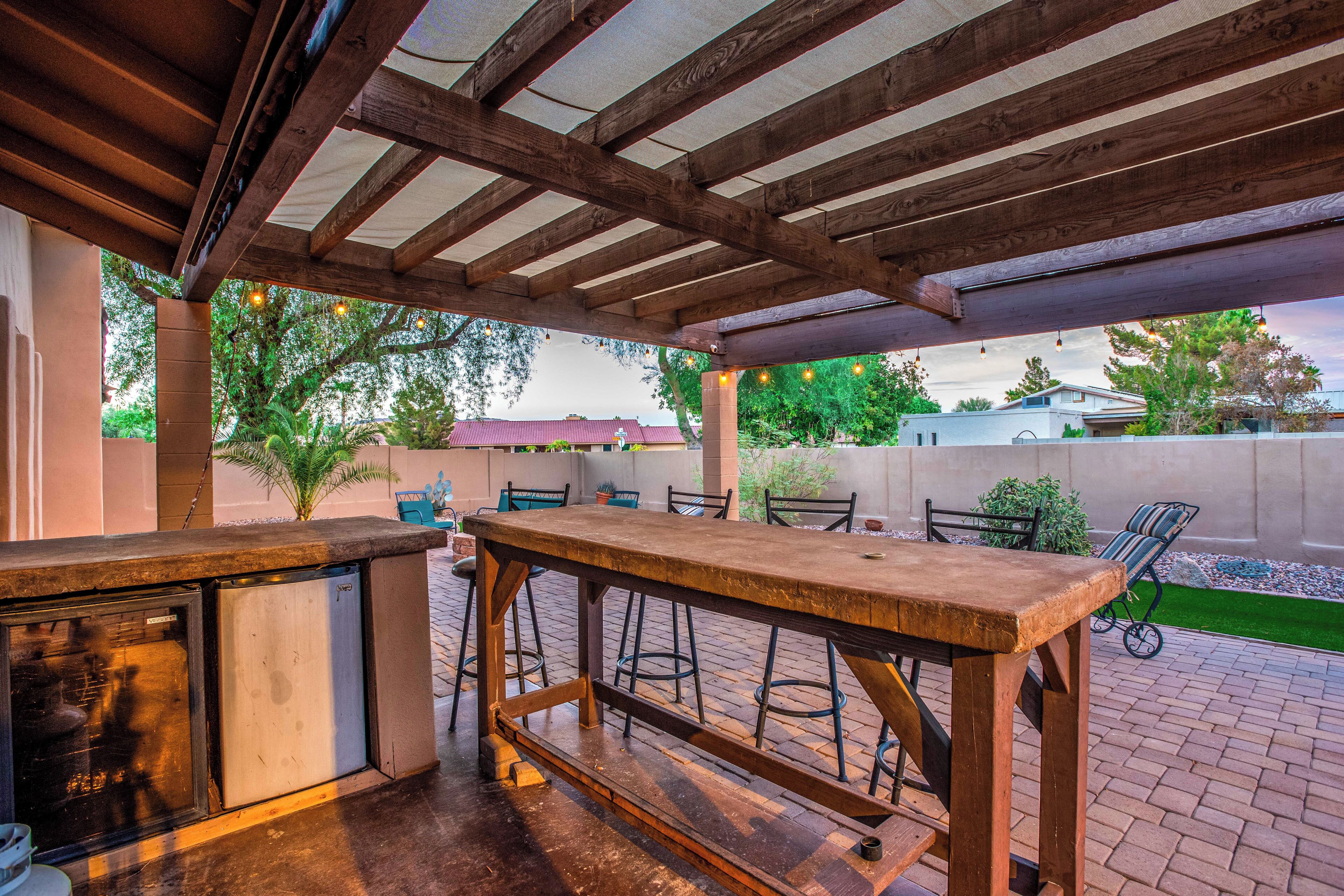Une terrasse extérieure avec une table en bois et des tabourets de bar, un barbecue encastré et des guirlandes lumineuses.