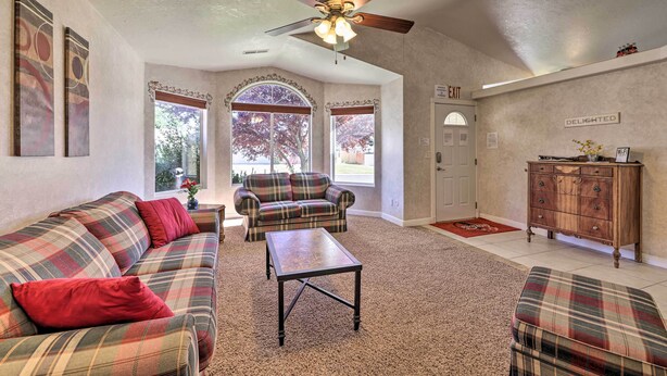 A living room with a plaid sofa, a coffee table, and a ceiling fan. There are two windows with decorative frames, a door labeled "EXIT," and a wooden cabinet. - Home w/ Game Room & Fire Pit: 30 Min to Zion! (La Verkin)