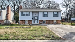 A two-story house with a garage door, brick foundation, and a front yard with grass and a driveway. - Maryland Vacation Home Near US Capital! (Riverdale)