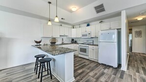 Uma cozinha moderna com armários brancos, bancada de granito com ilha, dois banquetas altas, eletrodomésticos de aço inoxidável e iluminação pendente.