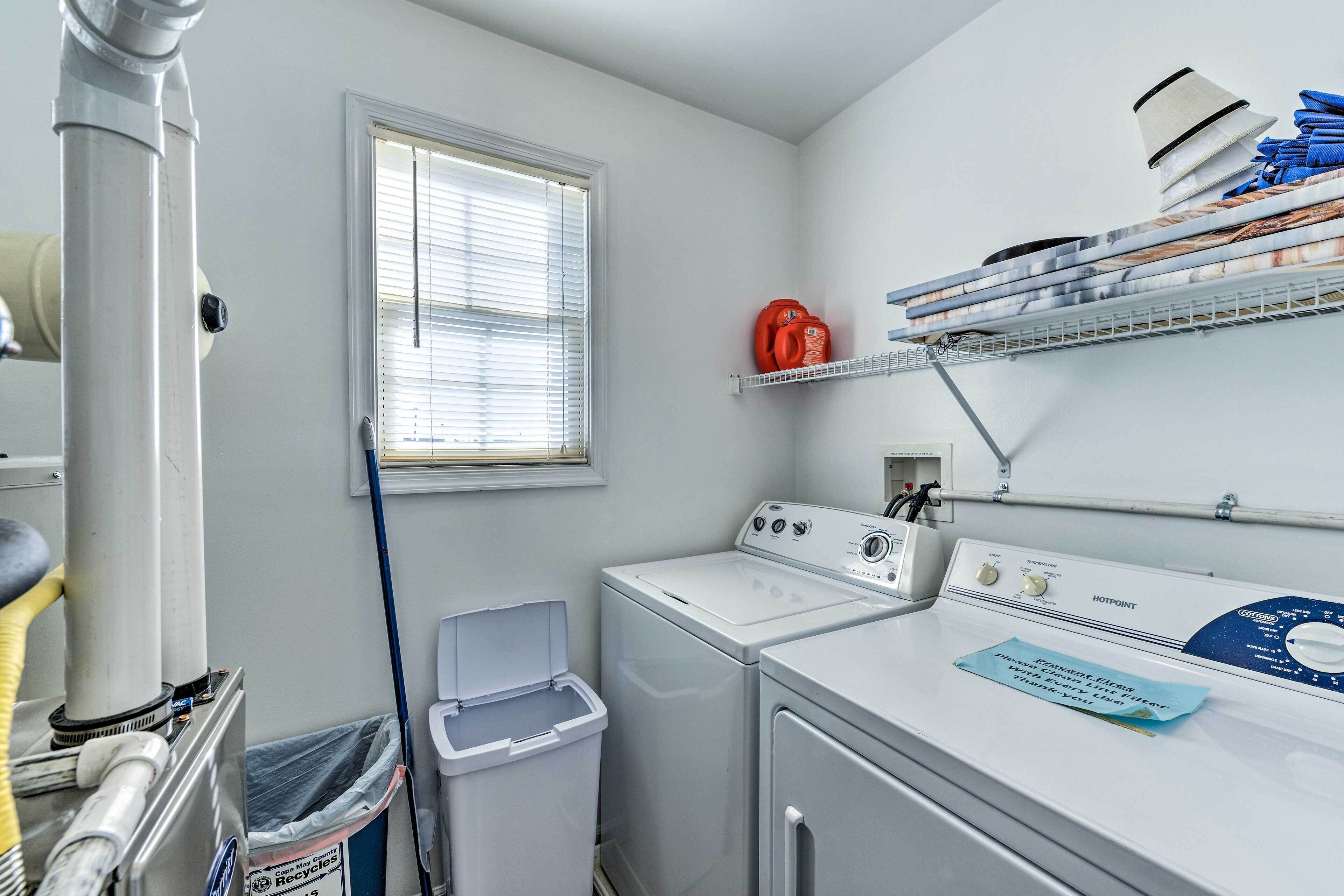 A laundry room with a front-loading washer and dryer, a window with blinds, a shelf with folded towels, and a trash can.