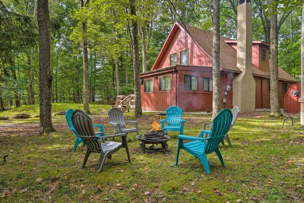 A cabin with a red exterior, surrounded by a forest. There are four Adirondack chairs and a small fire pit in the yard.