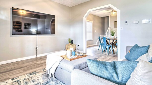 Een moderne woonkamer met een flatscreen televisie aan de muur, een gezellige zithoek met een bank en voetenbankje, en een eethoek met blauwe stoelen en een tafel.