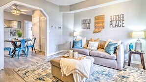 Een moderne woonkamer met een grijze bank, blauwe stoelen, een salontafel en een vloerkleed. Aan de muur hangen decoraties, waaronder een bord met 'Fountain Square' en een bord met 'Fletcher Place'.