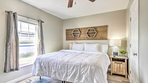 Een slaapkamer met een bed, een plafondventilator, een raam met gordijnen, een houten hoofdeinde, een nachtkastje met een lamp en een deur.