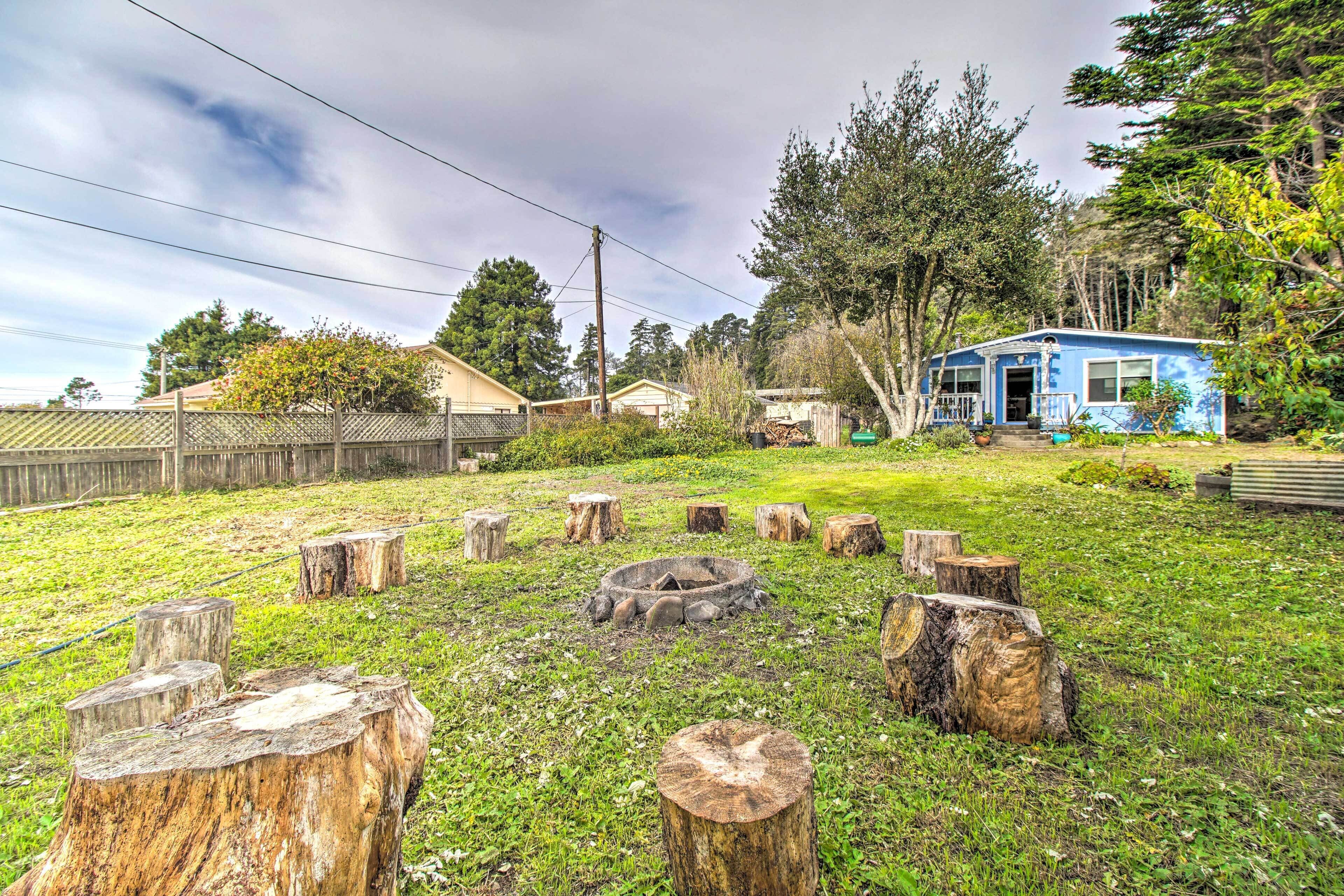 A backyard with a fire pit surrounded by tree stumps, a blue house, and a wooden fence.