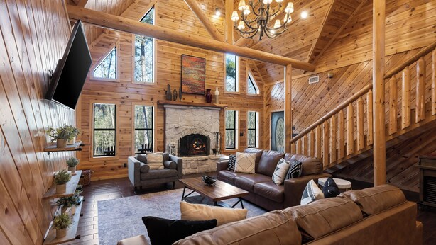 A log cabin interior with a stone fireplace, wooden walls, and a staircase. - Broken Bow Family Cabin w/ Private Hot Tub! (Broken Bow)