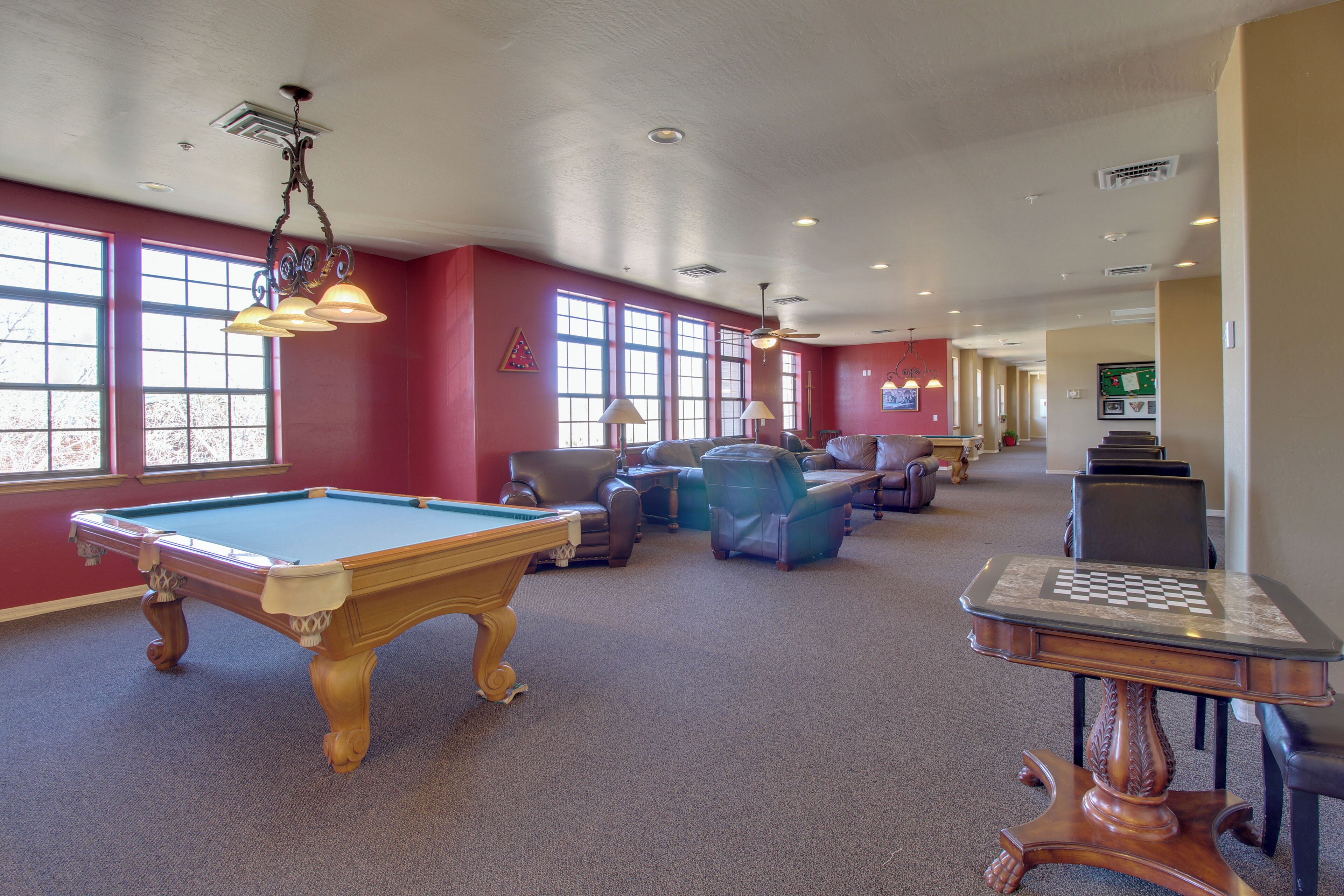 A pool table, lounge chairs, and a chess table in a room with large windows and red walls.